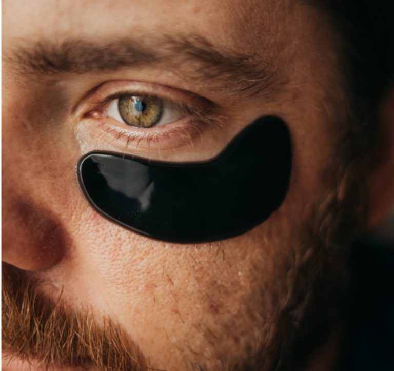 Side view of a bearded man with a black under eye gel mask under one eye. 