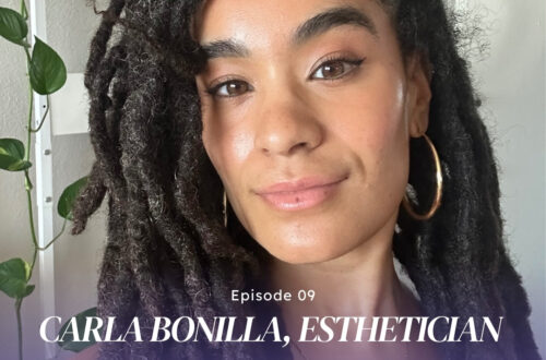 L.A.-based esthetician Carla Bonilla smiling into the camera with a green plant on a shelf behind her.